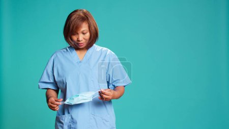 Photo for Tired nurse taking off protective face mask after finishing hospital work shift. Asian clinic employee pleased to finally remove stifling sanitary mask, isolated over blue studio background - Royalty Free Image