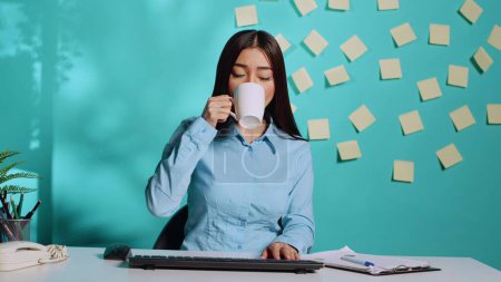 Téléchargez les photos : Femme d'affaires asiatique prenant une tasse de café lors d'une réunion de vidéoconférence en ligne, écoutant attentivement et bavardant à l'aide du clavier. Employé dans un bureau moderne sur fond bleu studio - en image libre de droit