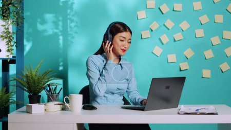 Téléchargez les photos : Heureuse femme asiatique profitant de la musique sur écouteurs, chantant et dansant au bureau créatif coloré tout en travaillant sur ordinateur portable. Femme d'affaires dans un lieu de travail détendu sur fond bleu studio - en image libre de droit