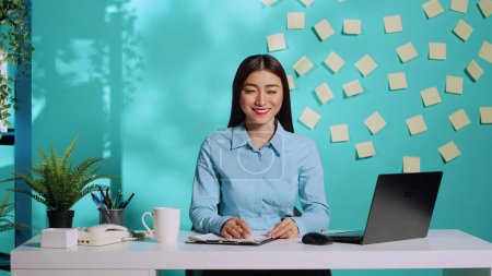 Foto de Feliz alegre mujer de negocios asiática sentada en el moderno escritorio de oficina creativa brillante sobre fondo de estudio azul. Empleado sonriente en un lugar de trabajo de administración relajado - Imagen libre de derechos
