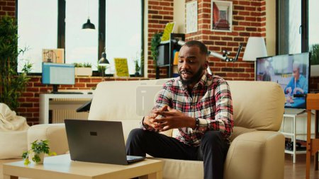 Photo for Smiling happy african american man excited to catch up with friends during online meeting while relaxing in stylish warm apartment. Person enjoying day off at home, chatting with friends in video call - Royalty Free Image