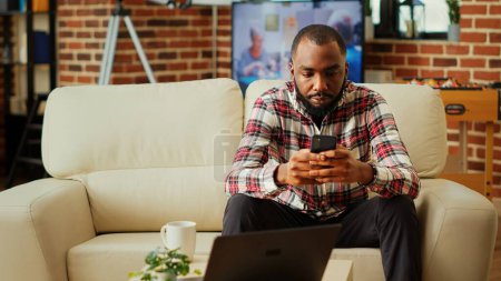 Photo for Annoyed african american man impatiently texting always late friend over supposed meeting hour. Person sitting on cozy sofa in modern stylish apartment living room typing on smartphone - Royalty Free Image