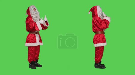 Photo for Saint nick in costume does thumbs up standing over full body greenscreen background, expressing like and agreement symbol in studio. Young man in santa suit shows approval sign. - Royalty Free Image