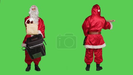 Photo for Deliveryman in red santa suit carrying backpack with pizza box and paper bag order, christmas eve delivery concept. Young man saint nick character against full body greenscreen backdrop. - Royalty Free Image