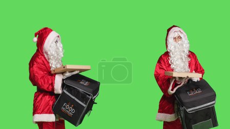 Foto de Deliveryman Santa Claus en carácter llevando bolsa térmica con comida rápida mientras celebra la víspera de Navidad en la pantalla verde. Hombre vestido con traje festivo de temporada con traje rojo y blanco. - Imagen libre de derechos