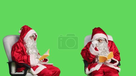 Téléchargez les photos : Père Noël lecture livre de fiction en studio, profiter de la littérature ou de la poésie conférence pour la connaissance. Saint Nick assis sur une chaise avec conte de fées à la main, fond d'écran vert. - en image libre de droit