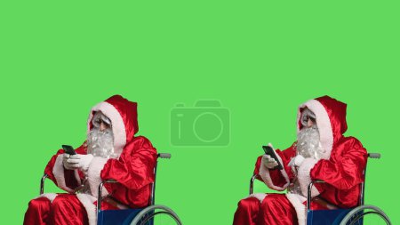 Photo for Saint nick texting and using videocall on smartphone, attend online teleconference while he is in a wheelchair dressed in red costume. Santa claus character with impairment works on phone. - Royalty Free Image