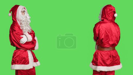Téléchargez les photos : Jeune homme confiant en costume traditionnel du Père Noël, avec barbe blanche et lunettes. Debout sur fond vert représente l'esprit de fête pendant la saison de Noël. - en image libre de droit