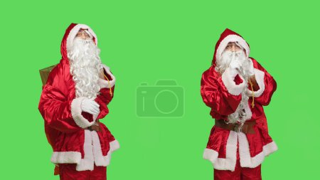 Photo for Father christmas shows mute symbol, espressing privacy and silence over greenscreen in studio. Santa claus with gifts bag doing hush sign to keep secret, private seasonal winter character. - Royalty Free Image