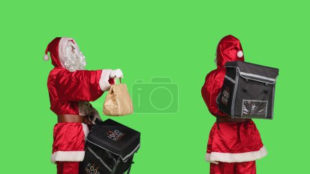 Photo for Festive man in santa claus suit deliver food with paper bag, posing with thermal backpack over greenscreen backdrop. Person in traditional holiday costume spreading christmas spirit. - Royalty Free Image