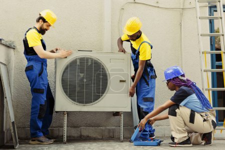 Seasoned mechanic coworkers installing outside air conditioner for customer. Adept repairmen commissioned to optimize new HVAC systems performance, ensuring it operates at maximum efficiency