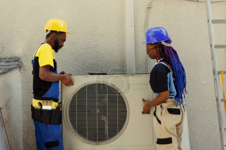 Téléchargez les photos : Des collègues d'ingénieurs afro-américains ouvrent un climatiseur rouillé hors service pour le remplacer par un nouveau condenseur extérieur performant après avoir vidangé le réfrigérant et remplacé les conduits - en image libre de droit