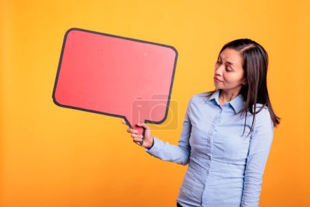 Photo for Asian young adult holding empty red speech bubble with copy space, advertising beauty product. Woman standing in studio over yellow background, looking at blank dialog frame with place for text - Royalty Free Image