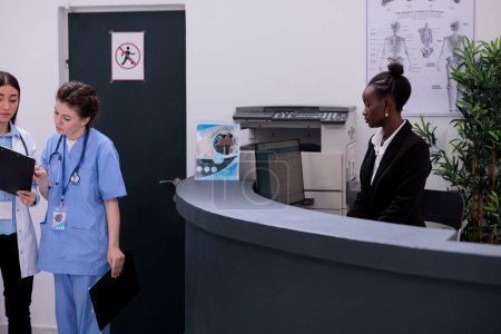 Téléchargez les photos : Réceptionniste afro-américain debout au comptoir de réception vérifiant la consultation médicale sur ordinateur lors de la visite de contrôle à l'hôpital. Fixation des rendez-vous de suivi pour les patients. - en image libre de droit