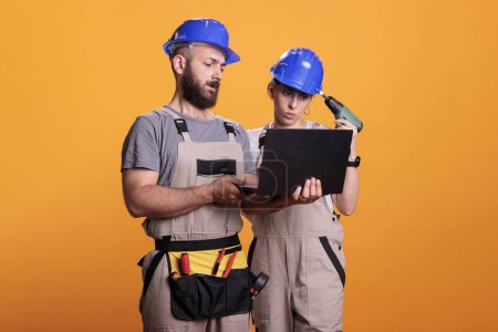 Photo for Professional builders in studio holding laptop, looking at building blueprint for new renovation project. Renovators wearing renovating uniform making decisions together as team. - Royalty Free Image