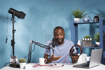 Photo for African american blogger reviewing headphones and recording video for internet channel. Social media influencer presenting device while streaming online and looking at camera in home studio - Royalty Free Image