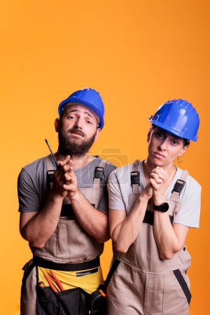 Photo for Religious repairman and woman praying to god for good projects, begging for help. Team of engineering experts holding prayer hands to ask for fortune and luck, wearing overalls and helmets. - Royalty Free Image