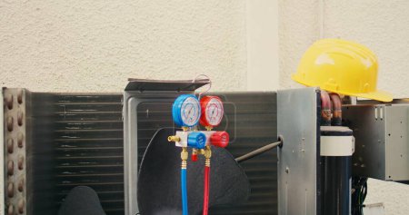 Photo for Close up of safety hardhat and pressure measurement device used for checking HVAC system refrigerant in need of maintenance. Set of freon levels benchmarking tool mounted on air conditioner - Royalty Free Image