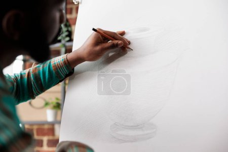Foto de Estudiante dibujando con lápiz sobre lienzo. Artista afroamericano que se mantiene enfocado mientras crea arte en estudio o taller, sentado en el caballete dibujando un jarrón básico usando lápiz - Imagen libre de derechos