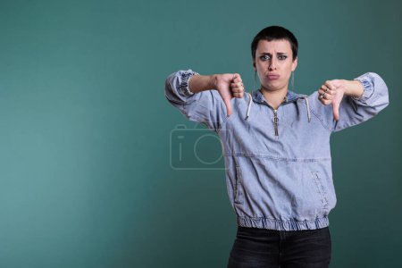 Photo for Serious disappointed woman doing thumbs down gesture while posing upset in studio over isolated background. Dissatisfied female giving disapproval sign gesturing negativity symbol. - Royalty Free Image
