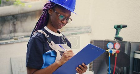 Photo for Portrait shot of smiling african american expert doing inspection of refrigerant levels and necessary repairs to prevent major breakdowns. Worker checking hvac system, writing findings on clipboard - Royalty Free Image