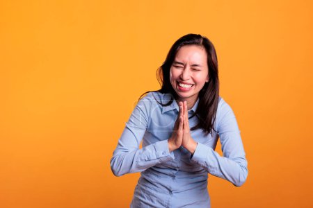 Téléchargez les photos : Femme philippine joyeuse priant Dieu pour le pardon, posant avec les paumes ensemble en studio sur fond jaune. Un jeune adulte plein d'espoir montrant un geste spirituel religieux - en image libre de droit