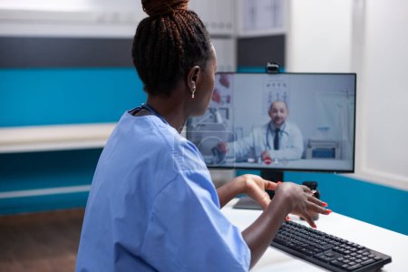 Photo for African american nurse at desk in a video meeting communicating with doctor coworker in a modern medical office. General practitioner in telehealth videocall with collegue - Royalty Free Image