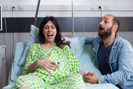 Foto de Mujer embarazada que tiene contracciones dolorosas, entrar en trabajo de parto en la sala de hospital, mientras que el marido nervioso gritando por el médico y la asistencia médica. Pareja joven caucásica preparándose para el parto - Imagen libre de derechos