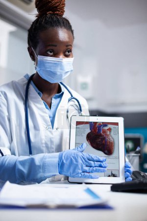 Photo for Cardiac specialist explaining heart informations to patient on tablet in doctors office. Cardiologist providing healthcare diagnosis wearing face mask and medical gloves in clinical consultation - Royalty Free Image