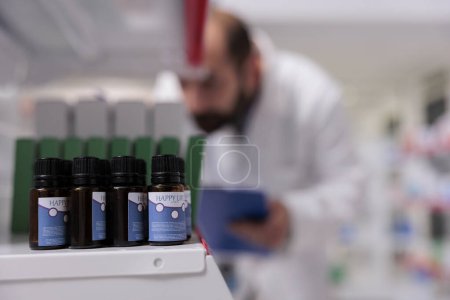 Photo for Selective focus of essential oil on pharmacy shelves is ready to be buy by client, man doing inventory in background. Drugstore is a vital resource for the community, providing access health services. - Royalty Free Image