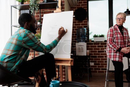 Foto de Joven chico afroamericano centrado en aprender a dibujar con el profesor, estudiante masculino sentado en el caballete en el aula de dibujo de objetos simples con lápiz, tomando clases de arte. Dibujo y auto-expresión - Imagen libre de derechos