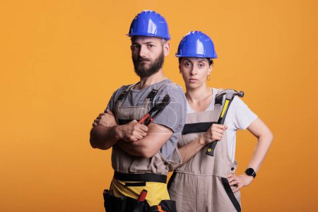 Serious construction workers posing with pair of pliers and hammer, holding slegdehammer and renovating tools in studio. Man and woman working as constructors with overalls and helmet.