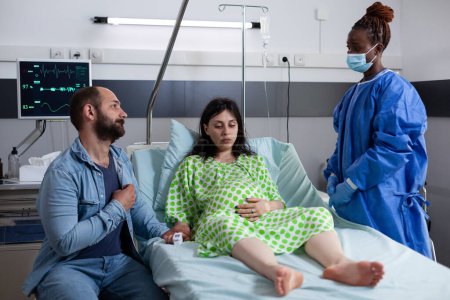 Téléchargez les photos : Chirurgien afro-américain discutant avec la femme enceinte avant l'opération, expliquant le processus de naissance dans la salle d'hôpital. Femme enceinte couchée au lit ayant des contractions réconfortantes par son mari - en image libre de droit