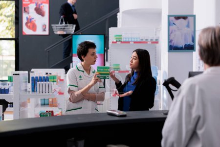 Foto de Drugstore customer complaining to pharmacist about purchased medicaments. Young asian woman and pharmaceutical assistant arguing about medical product price at pharmacy retail store - Imagen libre de derechos