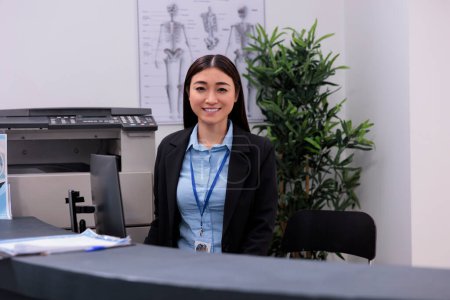 Photo for Smiling asian receptionist standing at hospital reception counter scheduling follow-up appointments for patients in waiting area. Worker using computer to record patient information. - Royalty Free Image
