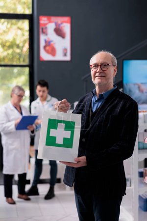 Photo for Senior man holding purchase shopping bag in drugstore portrait. Pharmacy elderly smiling customer buying prescription medicaments, standing in apothecary and looking at camera - Royalty Free Image