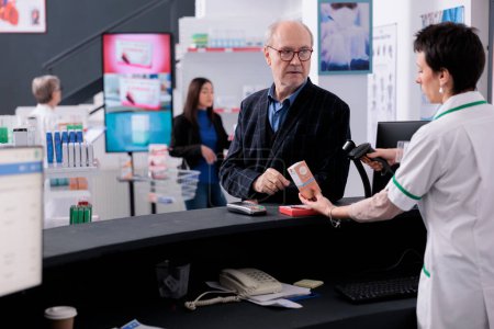 Téléchargez les photos : Elderly man giving pharmacy cashier medical product for scanning at checkout and discussing sunscreen with pharmaceutical worker. Customer visiting drugstore to buy sun protection lotion and vitamin - en image libre de droit