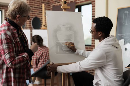 Foto de Young African American guy instructor teaching senior woman to draw during art class, teacher sitting at easel showing mature student how to hold and use pencil. Taking up drawing lesson in retirement - Imagen libre de derechos