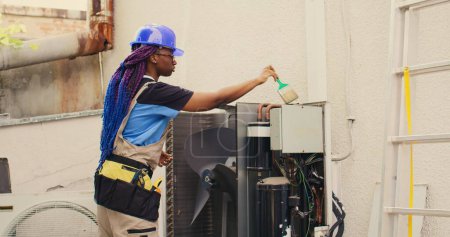 Foto de African american efficient mechanic commissioned by home owner for annual air conditioner routine maintenance, using soft dusting brush to clean dusty condenser coils and filters, outdoor - Imagen libre de derechos
