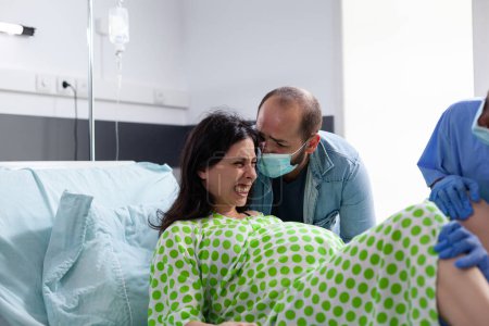 Téléchargez les photos : Hospital team helping pregnant woman to delivery baby in maternity clinic. Patient with pregnancy having painful contractions being comforting by husband while giving birth to child - en image libre de droit