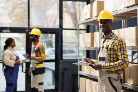 Photo for Shipment service warehouse manager checking orders list on clipboard portrait. Smiling young african american delivery operator working in storehouse and looking at camera - Royalty Free Image