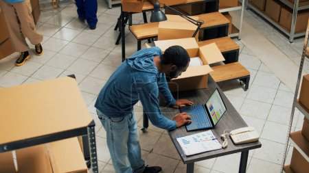 Foto de African american employee putting products in cardboard boxes, working on delivery and shipment. Young adult planning supply chain management, using laptop in warehouse storage room. - Imagen libre de derechos