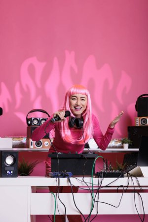 Photo for Musician standing at dj table holding microphone talking with fans while playing techno song using professional mixer console. Asian performer with pink hair enjoying to perform music at night in club - Royalty Free Image