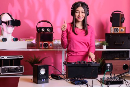 Photo for Positive musician standing at dj table doing thumbs up gesture while mixing electronic music and techno using mixer console. Asian artist playing stereo sounds with electronics and microphone - Royalty Free Image