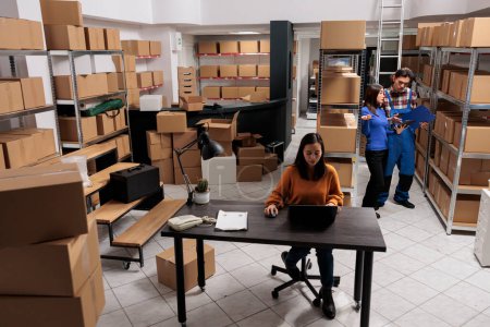 Photo for Warehouse asian employees team preparing order parcel for shipment in retail business storage room. Storehouse workers analyzing goods checklist and creating invoice on laptop - Royalty Free Image