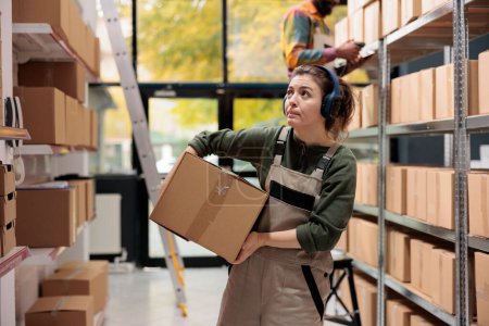 Photo for Storage room manager looking at shelves full with boxes, wearing headphones listening music while carrying cardboard box in warehouse. Employee working at customers orders preparing delivery - Royalty Free Image