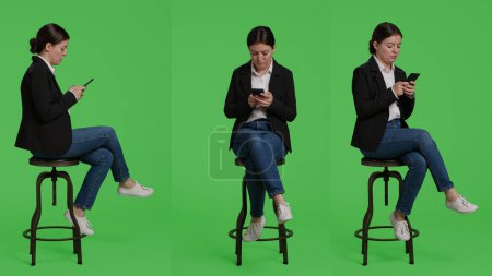 Photo for Modern female model browsing internet on smartphone, acting relaxed on chair in studio. Company employee in suit using mobile phone app to text messages, full body greenscreen backdrop. - Royalty Free Image
