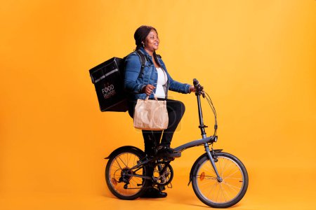 Photo for African american delivery woman holding paper bag while delivering lunch order to client using bike as transportation. Restaurant courier carrying takeout thermal backpack, takeaway service concept - Royalty Free Image