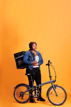 Photo for African american deliverywoman standing beside bicycle holding thermal backpack while delivering take out food to client during lunch time. Pizzeria courier employee carrying takeaway meal - Royalty Free Image
