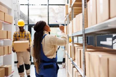 Photo for Industrial warehouse employee managing freight inventory, standing near shelf full of cartons. African american woman counting parcels and pointing with pen in shipment storehouse - Royalty Free Image